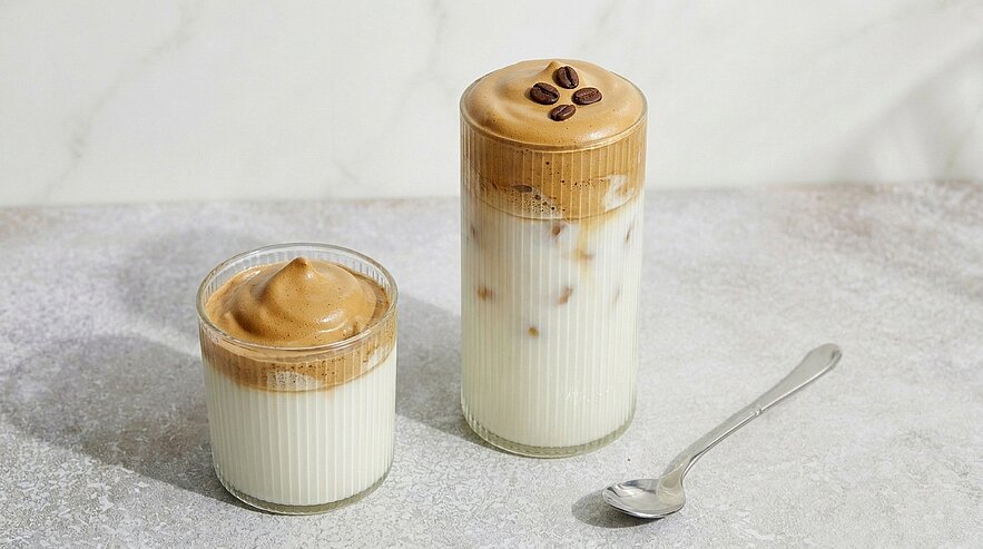 Zwei Gläser Dalgona-Kaffee: cremig aufgeschäumter, karamellfarbener Kaffeeschaum auf kalter Milch. Ein hohes Glas mit Eiswürfeln und drei Kaffeebohnen auf dem Schaum, daneben ein niedrigeres Glas mit dichtem Schaum.