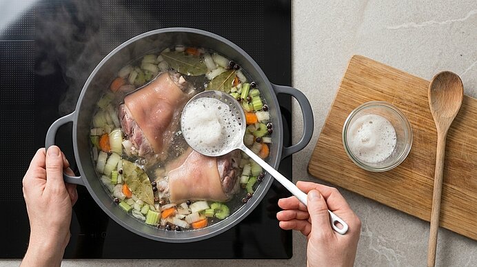 Topf mit kochender Brühe, Schweinehaxen, gewürfeltem Gemüse und Lorbeerblatt; Hände schöpfen Schaum mit Schaumlöffel, kleine Schüssel mit abgeschöpftem Schaum daneben.