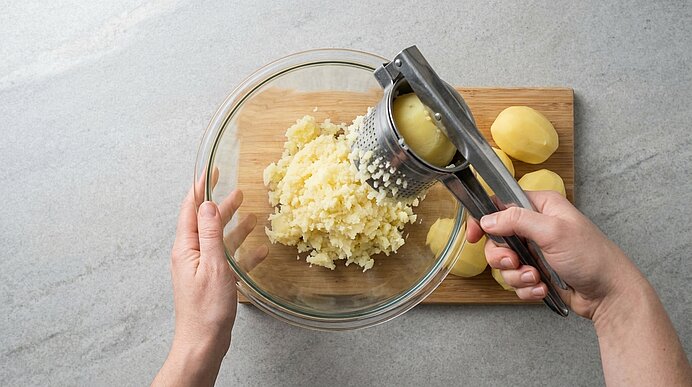 Zwei Hände pressen gekochte Kartoffeln durch eine Kartoffelpresse in eine Glasschüssel, daneben liegen geschälte Kartoffeln.