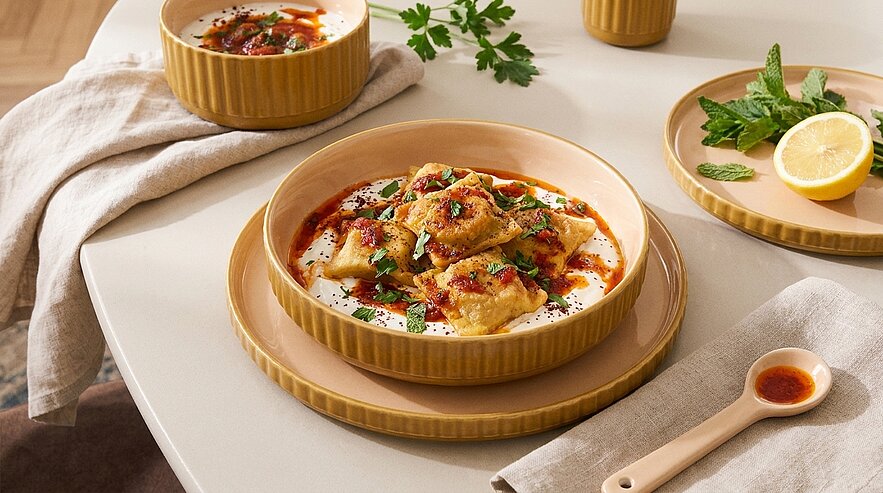 Schale mit gefüllten Teigtaschen (Ravioli) in cremiger Joghurt-/Sahnesauce, beträufelt mit Chiliöl und bestreut mit frischen Kräutern; daneben Teller mit Zitronenscheibe und Kräutern sowie ein Löffel mit Gewürz.