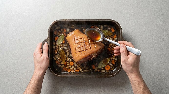 Zwei Hände begießen mit einem Löffel einen kreuzweise eingeschnittenen Braten in einem Bräter, umgeben von gewürfeltem Gemüse und Lorbeerblättern in Bratensaft.