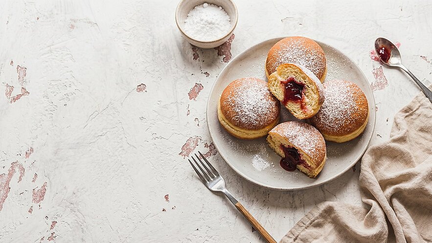 Auf einem Teller liegen mehrere gefüllte Donuts, von denen einer halbiert ist und eine rote Füllung zeigt. Die Oberfläche der Donuts ist mit Puderzucker bestreut. Daneben befindet sich eine Gabel und ein angerissener Serviettenstoff.