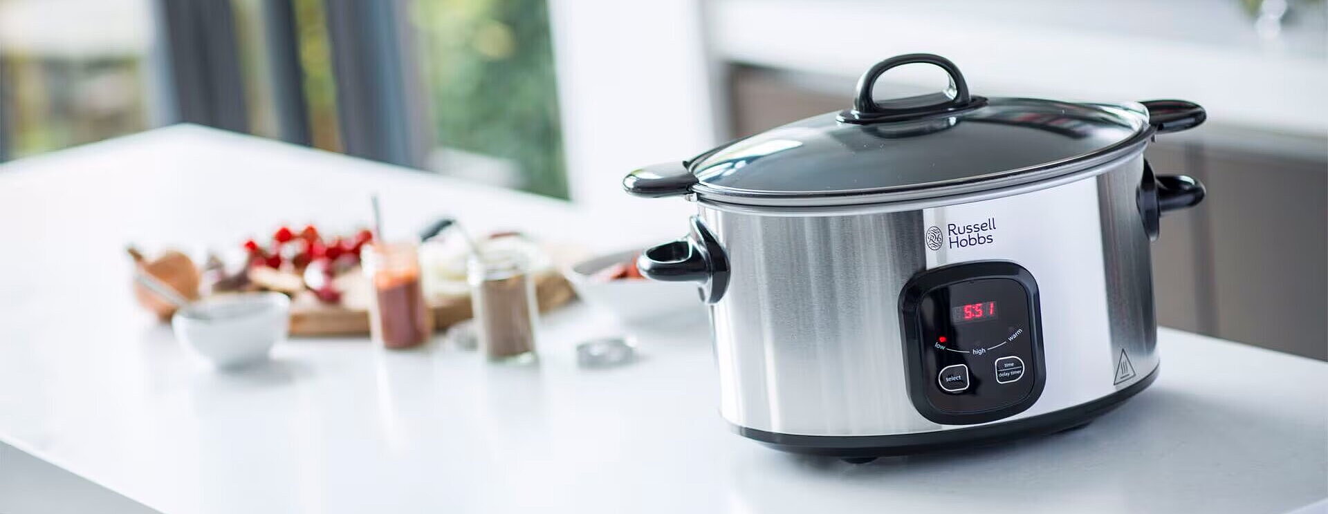Ein eleganter slow cooker von Russell Hobbs steht auf einer modernen, weißen Küchenarbeitsplatte. Er hat einen schwarzen Deckel und ein Bedienfeld mit digitalen Anzeigen. Im Hintergrund sind verschiedene Lebensmittel und Küchenutensilien zu sehen, die auf das Kochen vorbereiten.