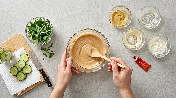 Zwei Hände verrühren Chili-Senf-Mayonnaise in einer Glasschüssel. Daneben stehen Schälchen, Rucola und Gurkenscheiben.