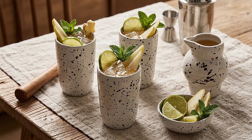 Drei gesprenkelte Keramikgläser mit Eis, Limetten- und Apfelscheiben sowie frischer Minze als Cocktailgarnitur auf rustikalem Tisch; daneben eine kleine Schale mit Limette und ein Kännchen.