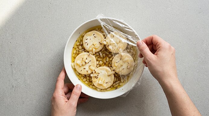 Hände spannen Frischhaltefolie über eine weiße Schüssel mit Brühe, klein gewürfelten Zwiebeln und vier runden, hellen Scheiben mit kreuzförmigen Einschnitten.