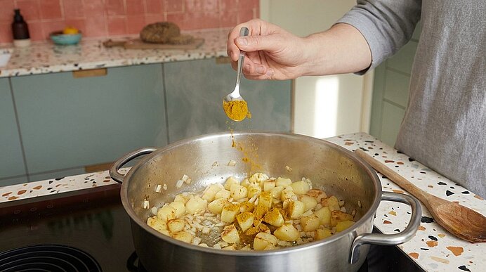 Hand einer Person streut mit einem Löffel Gewürz über eine Pfanne mit dampfenden, gewürfelten, sautierten Kartoffeln.