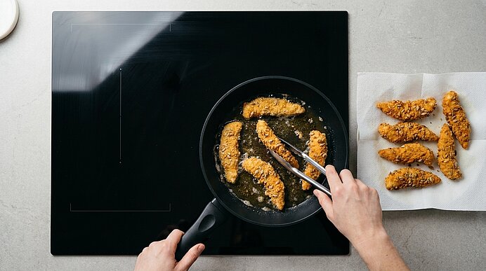 Obere Ansicht: Hände wenden panierte Hähnchenstreifen mit einer Zange in einer Pfanne; rechts auf Küchenpapier liegen bereits goldbraun gebratene Stücke.