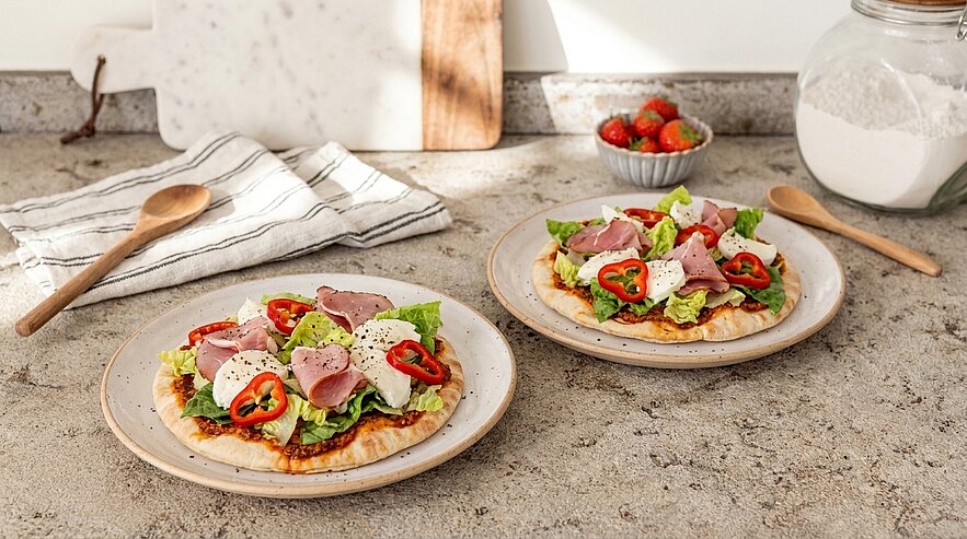 Zwei Teller mit belegten Fladenbroten auf einer Küchenarbeitsplatte: Salat, dünne Aufschnittscheiben, Käsewürfel und rote Paprikaringe.