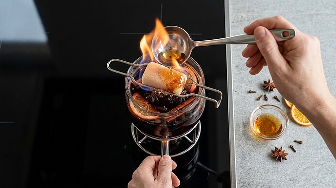 Hände flambieren einen Zuckerwürfel über einem Glas Glühwein: Mit einem Löffel wird Alkohol auf den Zucker gegossen und angezündet, sodass eine kleine Flamme über dem Getränk lodert.