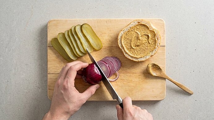 Zwei Hände schneiden eine rote Zwiebel auf einem Holzbrett neben Gewürzgurken und einer Semmelhälfte mit süßem Senf.