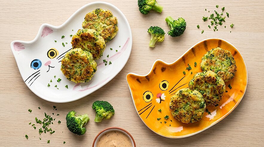 Zwei katzenförmige Teller, auf jedem drei goldbraune Brokkoli-Bratlinge; verstreute Brokkoliröschen und eine kleine Schale Dipsauce.