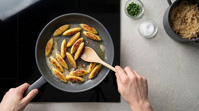 Zwei Hände braten goldbraune Schupfnudeln in einer Pfanne mit Butter; eine Hand rührt mit einem Holzspatel, neben der Pfanne stehen Sauerkraut, gehackte Kräuter und Salz.