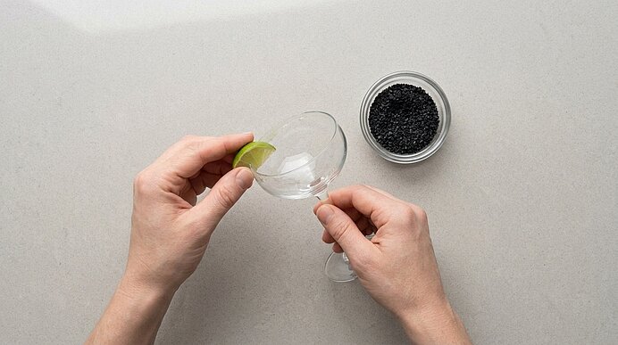 Zwei Hände reiben mit einer Limettenspalte den Rand eines Cocktailglases ein. Daneben steht eine Schale mit schwarzem Salz.