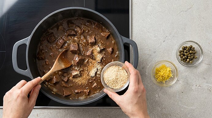 Zwei Hände rühren in einem Topf ein Ragout mit Fleischstücken in brauner Sauce; eine Hand streut Semmelbrösel, daneben Schälchen mit Zitronenschale und Kapern.