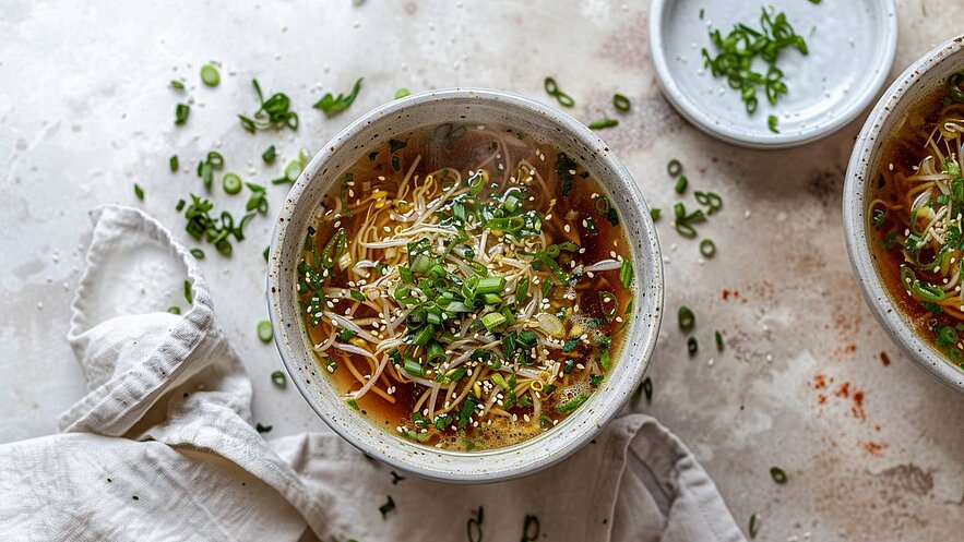 Eine Schüssel heiße Nudelsuppe ist mit frischen Kräutern, Sprossen und Sesam garniert. Die Suppe hat eine dunkelbraune Brühe und die Zutaten sind gut sichtbar. Auf einem hellen, strukturierten Untergrund liegen einige geschnittene Frühlingszwiebeln und ein Tuch.