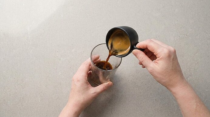 Zwei Hände gießen frisch gebrühten Kaffee aus einer kleinen Kanne in ein klares Glas.