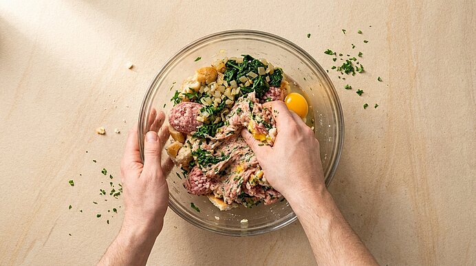 Zwei Hände vermengen in einer Glasschüssel rohes Hackfleisch mit Brotstücken, Ei und gehackten Kräutern.