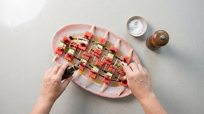 Zwei Hände beträufeln Wassermelone-Feta-Spieße auf einer Platte von oben gesehen mit Balsamico-Creme.