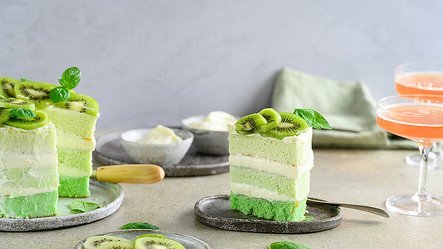 Ein Stück grüner Kuchen mit mehreren Schichten, dekoriert mit Kiwis und Minzblättern. Neben dem Kuchen befinden sich kleine Schalen mit Sahne und ein Löffel. Im Hintergrund stehen zwei Gläser mit einem rosa Getränk.