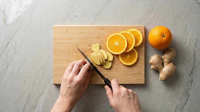 Zwei Hände schneiden auf einem Holzbrett dünne Ingwerscheiben; daneben liegen aufgeschnittene Orangen, eine ganze Orange und ein Stück Ingwer.