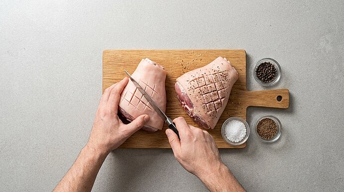 Zwei Schweinefleischstücke mit eingeschnittenem Fettrand auf einem Holzbrett; Hände machen Kreuzschnitte, daneben Schälchen mit Salz, Pfeffer und Gewürzen.