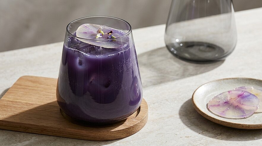 Glas mit violettem, mit Eis gefülltem Getränk, garniert mit einer essbaren Blüte, steht auf einem Holzuntersetzer auf hellem Tisch.