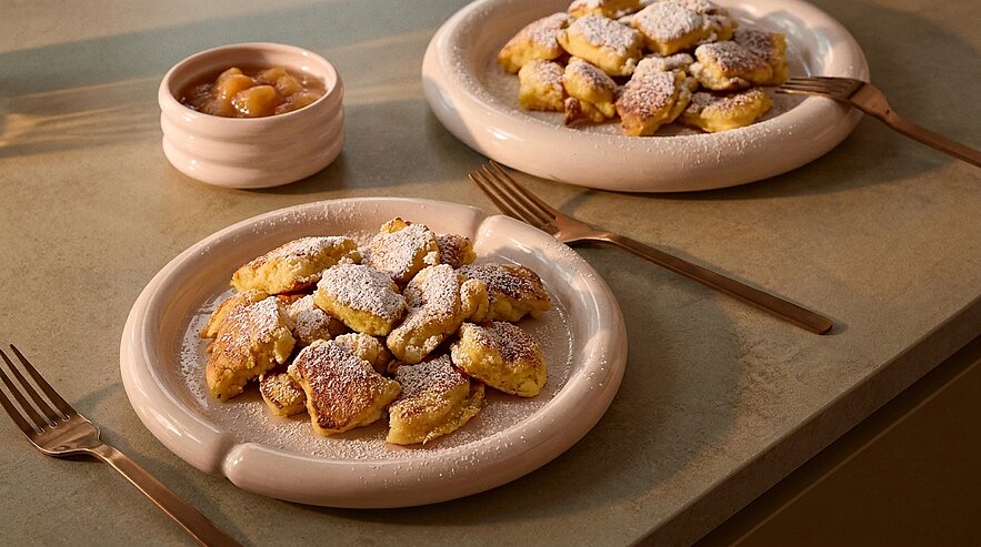 Zwei Teller mit kleinen, goldbraunen Pfannkuchenstücken (Kaiserschmarrn), bestäubt mit Puderzucker; daneben ein kleines Schälchen Fruchtkompott und Besteck.