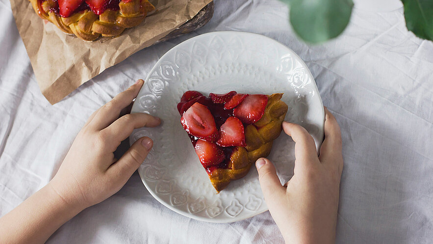 Ein Teller mit einem Stück Erdbeertorte wird von zwei Händen gehalten. Die Torte hat eine goldene Kruste und ist mit frischen Erdbeeren und Glanzüberzug dekoriert. Im Hintergrund ist eine weitere Torte auf einem Papier sichtbar.