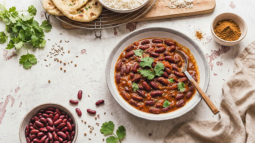 Eine Schüssel mit rotem Bohnen-Curry, garniert mit frischem Koriander, steht im Vordergrund. Daneben liegt eine Schale mit roten Bohnen. Im Hintergrund sind frisch gebackenes Naan-Brot und eine Portion Reis. Gewürze sind ebenfalls sichtbar.