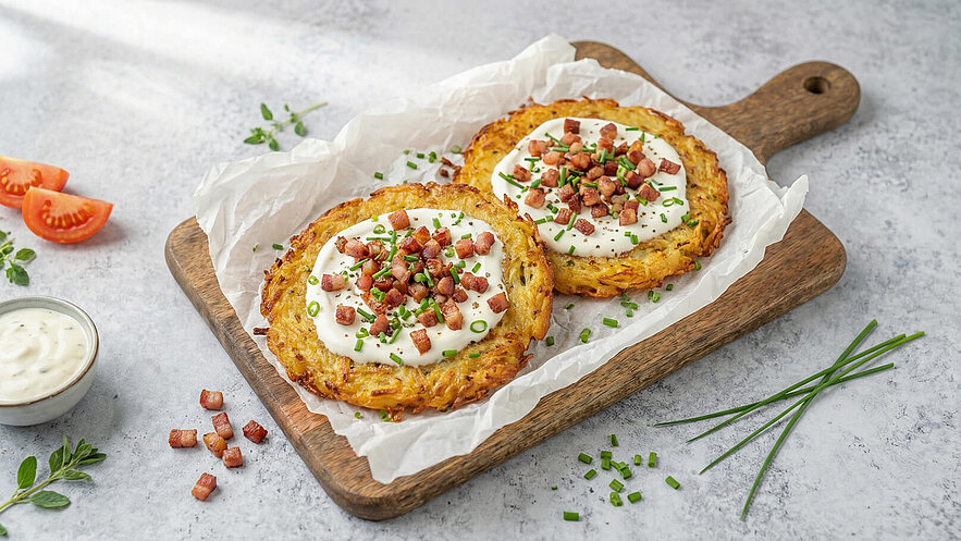 Auf einem Holzbrett liegen zwei goldbraune Kartoffelpuffer, garniert mit Sauerrahm, gebratenen Speckstückchen und feingehacktem Schnittlauch. Daneben befindet sich eine kleine Schüssel mit einer cremigen Sauce und einige frische Tomatenscheiben.