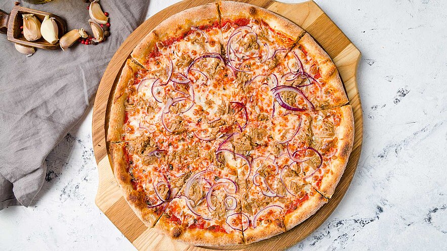 Eine frisch gebackene Pizza liegt auf einem Holzbrett. Sie ist mit Käse, roten Zwiebeln und Fleisch belegt und hat einen goldbraunen Rand. Die Pizza ist in Stücke geschnitten und befindet sich auf einem hellen Tisch.
