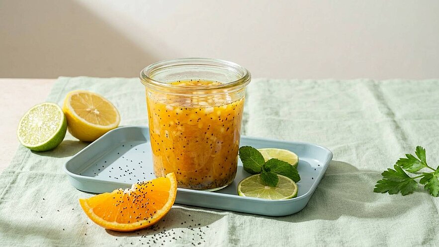 Ein Glas mit einer bunten Mischung aus Mango und Limette steht auf einem blauen Tablett. Daneben liegen eine orange und eine halbe Limette. Frische Minze und ein wenig Koriander ergänzen die Szene. Der Hintergrund ist hell und neutral gehalten.