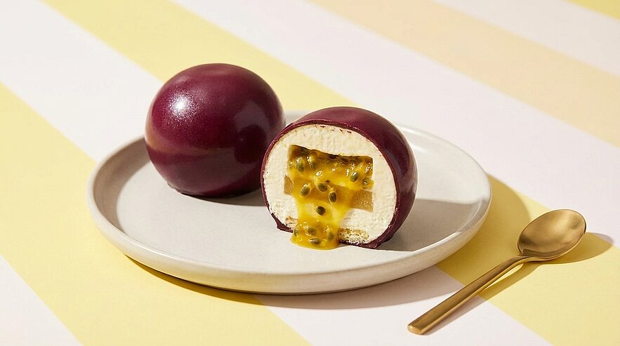 Zwei glänzende, als Passionsfrucht geformte Desserts auf einem Teller; eines halbiert, zeigt weiße Cremefüllung mit hervorquellendem Passionsfrucht-Püree und schwarzen Kernen; goldener Dessertlöffel daneben.