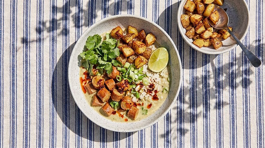 Schüssel mit cremiger Suppe oder Brei, garniert mit gebratenen Würstchenwürfeln, Frühlingszwiebeln, Koriander, Limettenscheibe und Chiliflocken; daneben kleine Schale mit gewürzten Bratkartoffeln.