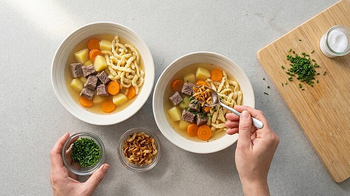 Zwei Schalen mit Suppe (Rindfleischwürfel, Kartoffelstücke, Karottenscheiben und Spätzle); Hände garnieren eine Schale mit Röstzwiebeln und Schnittlauch.