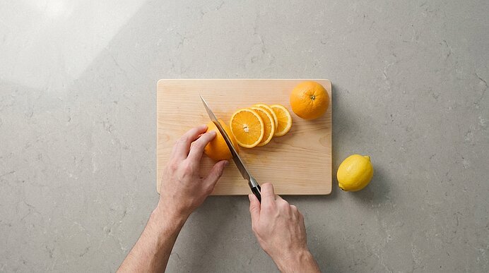 Zwei Hände schneiden eine Orange mit einem Küchenmesser auf einem Holzbrett; mehrere Orangenscheiben liegen daneben, ebenso eine ganze Orange und eine Zitrone.