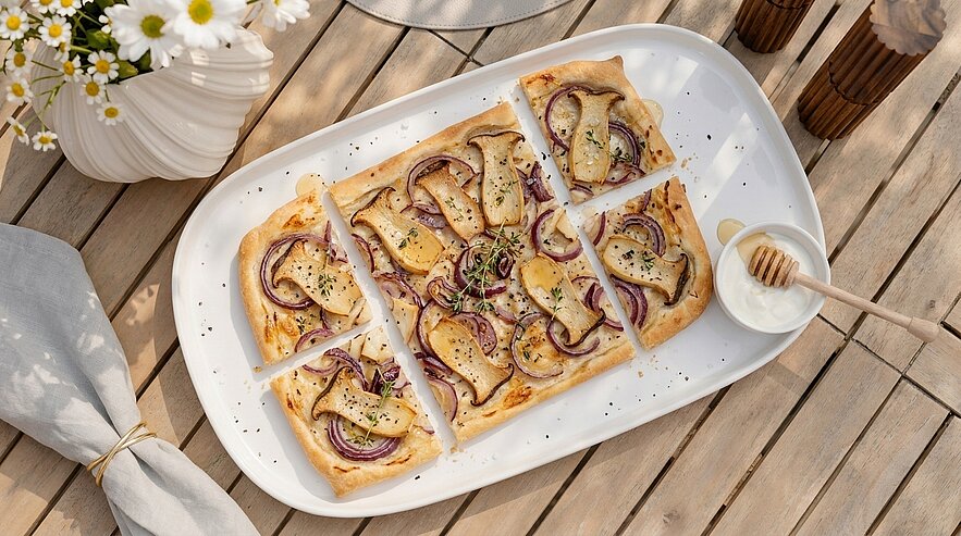 Rechteckiger Flammkuchen/Tarte mit dünn geschnittenen Birnen, roten Zwiebeln, geschmolzenem Käse und Kräutern, in sechs Stücke geschnitten auf einem weißen Servierteller; daneben ein Schälchen mit Honiglöffel.