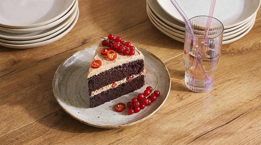 Ein Stück Schokoladenkuchen auf einem Teller, garniert mit roten Johannisbeeren und Orangenscheiben. Daneben steht ein Glas Wasser mit zwei Trinkhalmen. Im Hintergrund sind stapelbare, helle Teller sichtbar. Der Tisch hat eine Holzoberfläche.