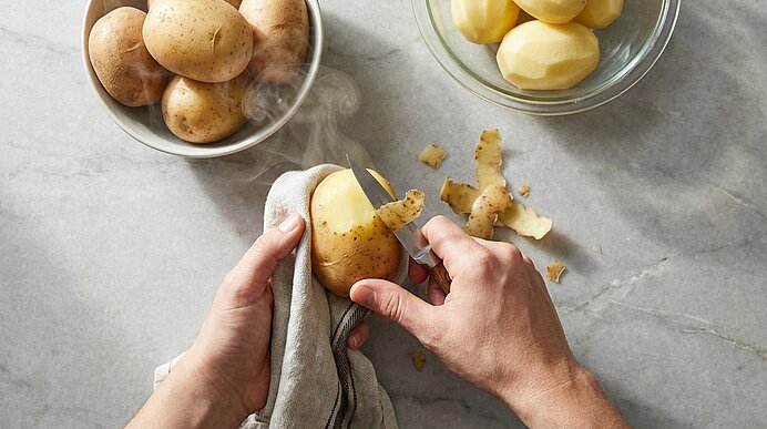 Nahaufnahme von Händen, die eine gekochte Kartoffel mit einem Messer über einem Tuch schälen; daneben Schüsseln mit ungeschälten und geschälten Kartoffeln sowie Kartoffelschalen.