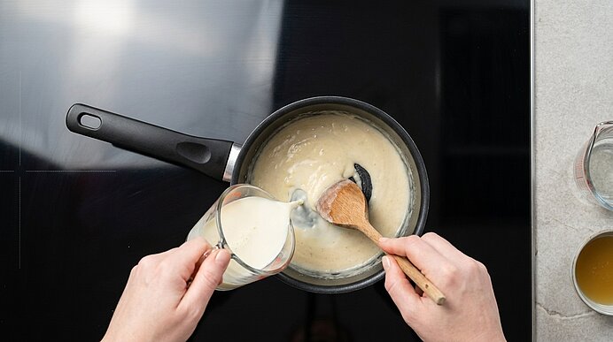 Zwei Hände rühren mit einem Holzlöffel eine helle, cremige Sauce in einem Topf, während aus einem Messbecher Milch in die Sauce gegossen wird.