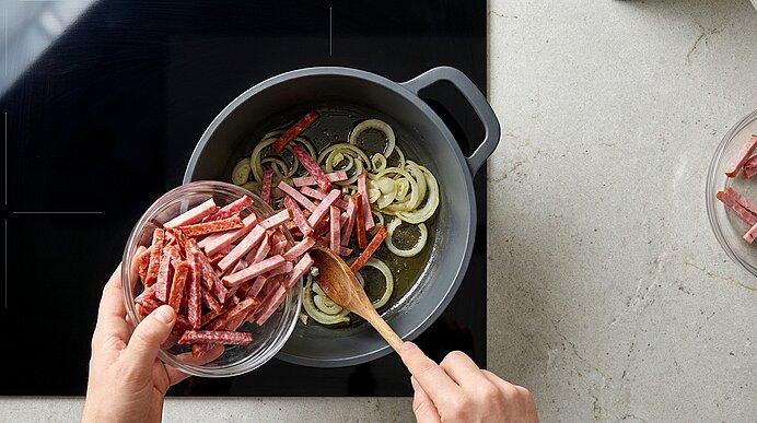 Hände geben Streifen Salami aus einer Glasschüssel in eine Pfanne mit angeschwitzten Zwiebelringen auf dem Herd; mit Holzlöffel wird gerührt.