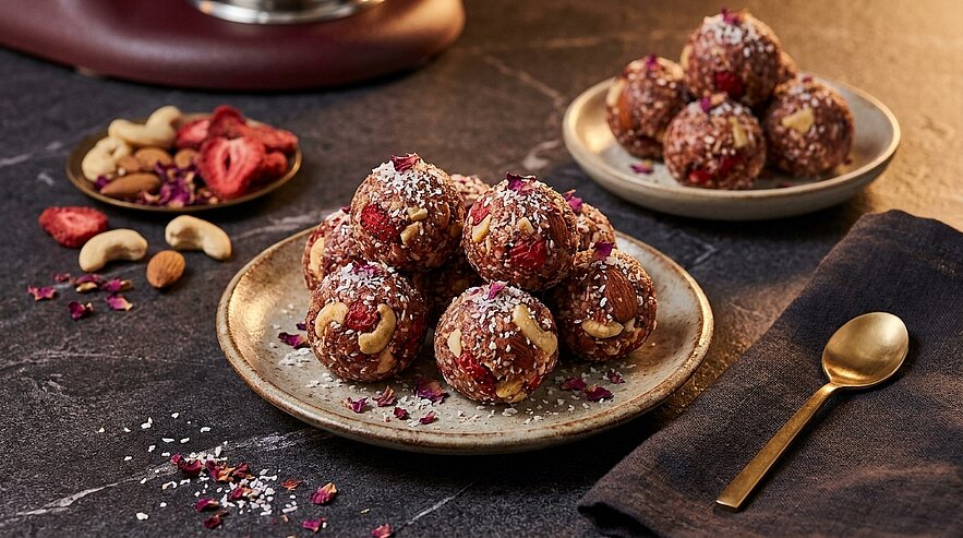 Mehrere kugelige Energiebällchen mit Nüssen und getrockneten Beeren, bestreut mit Kokosraspeln und Rosenblüten, auf zwei Keramiktellern angerichtet; daneben verstreute Cashewkerne.