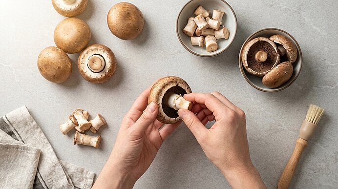 Zwei Hände drehen vorsichtig den Stiel aus einem braunen Champignon über einer hellgrauen Arbeitsplatte.