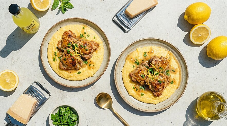 Zwei Teller mit cremiger Polenta, darauf gebratene Hähnchenschenkel, garniert mit Zitronenzesten und gehackter Petersilie.