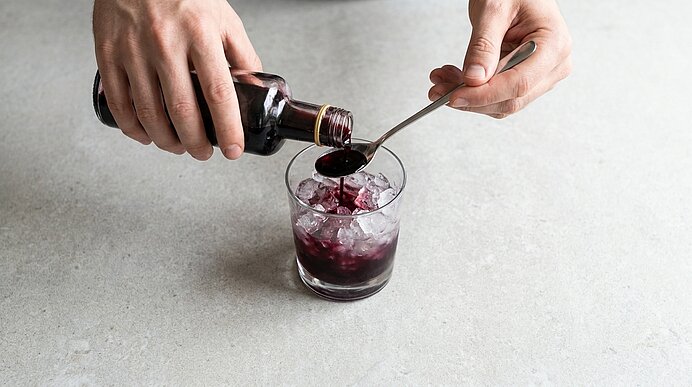 Hände gießen dunklen Sirup aus einer Flasche auf einen Löffel über einem Glas mit Eiswürfeln.