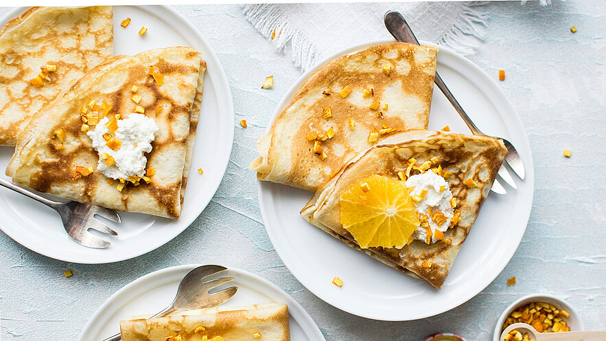 Zwei Teller mit frisch zubereiteten Crêpes sind sichtbar. Eine Crêpe ist gefüllt mit einer weißen Creme und dekoriert mit einer Orangenscheibe und zerstoßenen Mandarinen. Die Teller sind auf einem hellen Tischdeckchen platziert, umgeben von kleinen orangefarbenen Stücken.