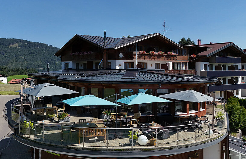 Ein modernes Gebäude mit einem runden, überdachten Außenbereich. Es sind Tische und Stühle unter blauen und grauen Sonnenschirmen aufgestellt. Im Hintergrund sind grüne Hügel und ein blauer Himmel zu sehen, was eine einladende, ruhige Atmosphäre vermittelt.