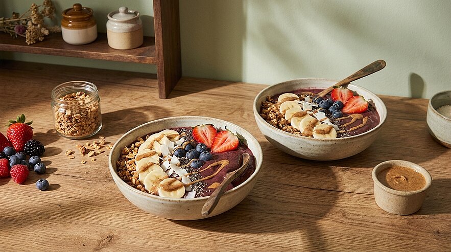 Zwei Smoothie-Bowls auf einer Holzfläche mit Löffeln, garniert mit Bananenscheiben, Erdbeeren, Blaubeeren, Granola und Kokosflocken.
