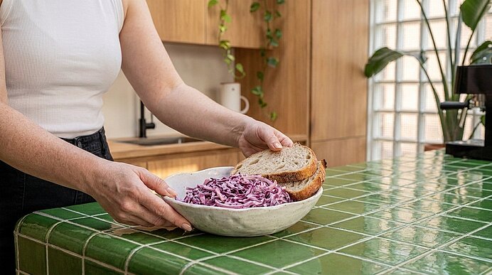 Hände einer Person stellen eine Schüssel mit Rotkohlsalat und zwei Brotscheiben auf eine grün gekachelte Küchenarbeitsfläche.